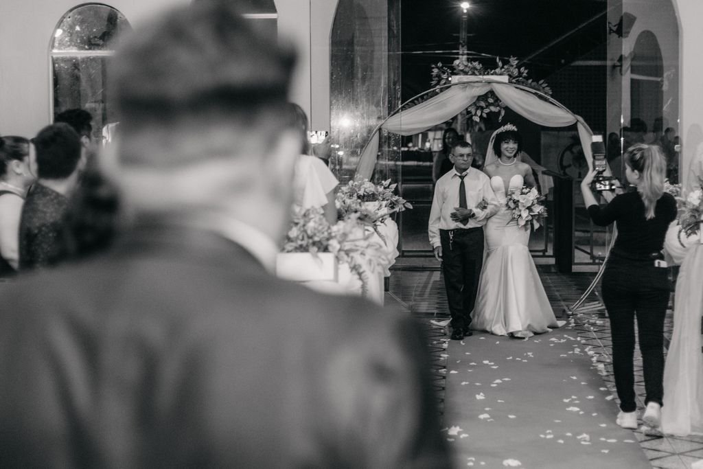 Father walks smiling bride down the aisle at indoor wedding ceremony. Captured in black and white.