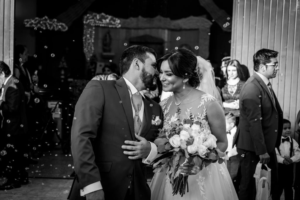 Joyful bride and groom exit their wedding ceremony surrounded by bubbles, holding a bouquet.